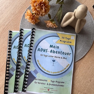 Könnte beinhalten: Drei spiralgebundene Bücher mit blau-grünen Umschlägen mit dem Text "Mein ADHS-Abenteuer". Die Bücher werden von orangefarbenen Blumen und einer kleinen beigefarbenen Figur auf einem grauen Platzdeckchen begleitet.