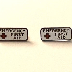 May include: Two rectangular enamel pins featuring a white background with a dark border. Each pin displays the text EMERGENCY FIRST AID in black capital letters, accompanied by a small red cross symbol positioned to the left of the text.