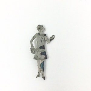 May include: A flat, silver-toned metal brooch shaped like a standing figure wearing a short dress and a headpiece. The metal surface shows signs of oxidation and wear, with dark and blue-green patina spots visible across the body and limbs.