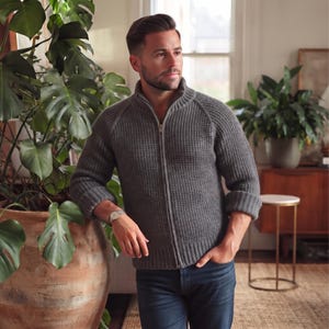 May include: A charcoal gray, long-sleeved, full-zip sweater featuring a textured waffle-knit pattern, a high collar, and ribbed cuffs. The garment is worn with dark blue denim jeans and a silver-toned wristwatch on the left wrist.