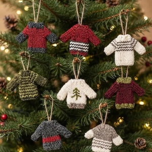 May include: A collection of eight miniature knitted sweater ornaments hanging from strings. The sweaters feature various designs including solid colors, horizontal stripes, and a small green tree pattern in red, white, gray, green, and burgundy yarn.