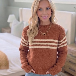 Può includere: Un maglione in maglia color ruggine con scollo rotondo e due strisce orizzontali color crema sul petto e sulle maniche. Il maglione presenta maniche lunghe con polsini a coste e un orlo a coste, abbinato a una collana a catena dorata.