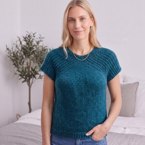 May include: A short-sleeved, teal-colored knitted top featuring a round neckline and a textured ribbed pattern across the upper chest and shoulders. The garment has a straight hemline and a smooth, stockinette stitch finish on the main body.