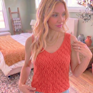 May include: A sleeveless coral-colored knit top featuring a repeating leaf-like textured pattern throughout the fabric, a V-neckline, and ribbed trim along the collar and arm openings, paired with a layered gold chain necklace.