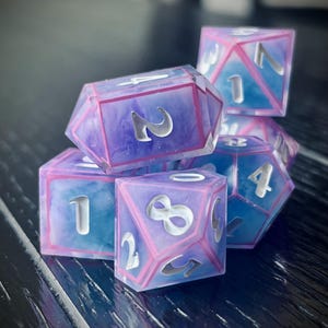 May include: A set of polyhedral dice with a translucent design. The dice feature a mix of blue and purple hues with pink borders and silver numbering. These dice are designed for tabletop gaming.