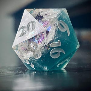 May include: A handcrafted, twenty-sided die with a clear, teal, and white design. The die features white numerals and internal iridescent accents. The numbers 20, 9, 16, and 8 are visible. This unique die is suitable for tabletop gaming.
