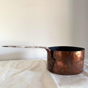 May include: A round copper saucepan with a long, flat handle attached to the side by a triangular plate with three visible rivets. The exterior surface of the pan shows a weathered, metallic finish with variations in color and texture.