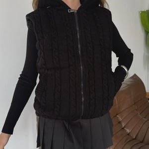 Puede incluir: Chaleco negro de punto trenzado con cremallera y capucha. El chaleco se lleva sobre una camiseta negra de manga larga y se combina con una falda plisada negra. Se ve una pulsera plateada en la mu&ntilde;eca.