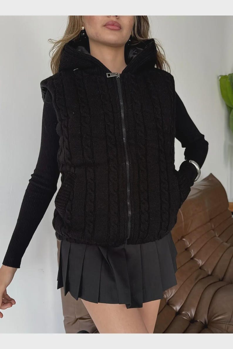 Puede incluir: Chaleco negro de punto trenzado con cremallera y capucha. El chaleco se lleva sobre una camiseta negra de manga larga y se combina con una falda plisada negra. Se ve una pulsera plateada en la mu&ntilde;eca.