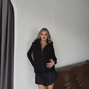 Puede incluir: Un chaleco negro con capucha y cierre de cremallera, combinado con una camiseta negra de manga larga y una falda plisada negra. El conjunto se completa con botas negras. El chaleco tiene un dise&ntilde;o de punto trenzado.