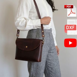 Könnte beinhalten: Dunkelbraune Leder-Umhängetasche mit Klappenverschluss und verstellbarem Riemen. Die Tasche hat einen messingfarbenen Knopfverschluss und wird über der Schulter getragen. Die Tasche ist ca. 25 cm breit und 20 cm hoch.