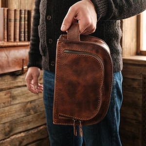 Peut inclure: Un sac de voyage en cuir marron avec une poche zippée incurvée et une poignée. Le sac a une surface texturée et une fermeture éclair. Le sac est tenu par une personne portant un pull sombre et un jean bleu.