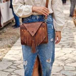 May include: A brown leather crossbody bag with fringe and a flap closure. The bag is worn with a denim skirt and a cream-colored blouse. The bag has a long strap and is adorned with gold-colored studs.