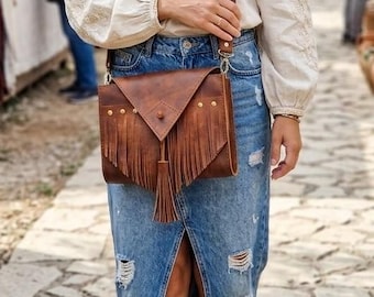 May include: A brown leather crossbody bag with fringe and a flap closure. The bag is worn with a denim skirt and a cream-colored blouse. The bag has a long strap and is adorned with gold-colored studs.