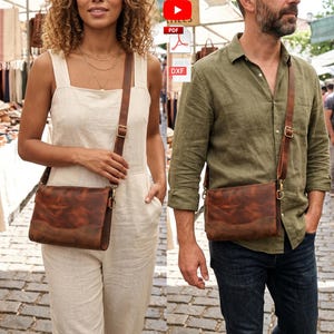 Può includere: Una borsa a tracolla rettangolare in pelle marrone invecchiata con una sezione superiore liscia e un pannello inferiore testurizzato più scuro. La borsa presenta una sottile tracolla in pelle regolabile con fibbia e finiture in metallo ottonato.