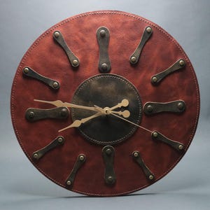 Puede incluir: Reloj de pared redondo con esfera de cuero marrón rojizo y centro de color bronce oscuro. El reloj presenta manecillas e indicadores de hora de color bronce. El diseño del reloj tiene una estética vintage e industrial.