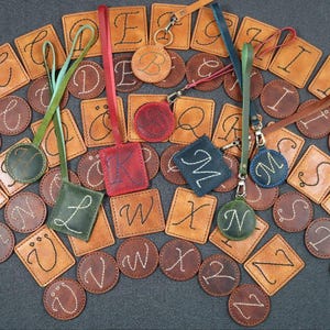 以下が含まれることがあります： アルファベットの文字がそれぞれ付いた革製のキーホルダーとタグのコレクション。キーホルダーは正方形や円形など様々な形で、ステッチで文字が施されています。一部には色のアクセントとストラップが付いています。