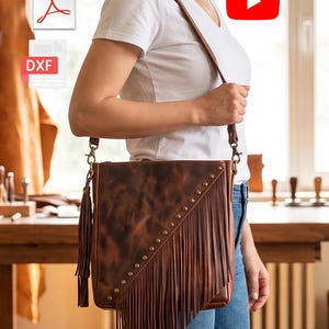 Puede incluir: Un bolso bandolera de cuero marrón con una fila diagonal de tachuelas metálicas color latón y flecos largos. El bolso incluye una borla de cuero a juego en el lateral y una correa de hombro fina y ajustable con clips de metal.
