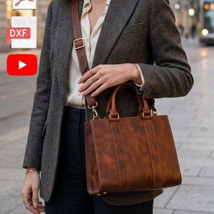 May include: A brown leather handbag with a top handle and shoulder strap. The bag has a rectangular shape with vertical stitching details. The person is wearing a gray blazer, a white blouse, and dark blue pants.