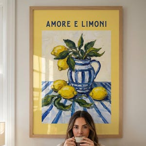May include: A framed painting with the words "AMORE E LIMONI" features a still life of lemons, a blue and white vase with greenery, and a yellow background. A person is holding a cup of coffee in front of the painting.