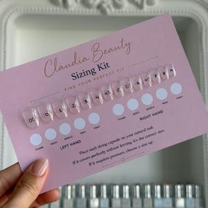 Op de afbeelding: Een roze Claudia Beauty Sizing Kit kaart met doorzichtige nagelmaat capsules. De kaart heeft de tekst "Find Your Perfect Fit" en gebruiksaanwijzingen. De kaart wordt door een hand vastgehouden.