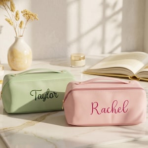 Puede incluir: Dos neceseres rectangulares con asas superiores y cremalleras doradas. Uno es de color verde claro con el nombre Taylor bordado en cursiva verde oscuro, y el otro es de color rosa claro con el nombre Rachel bordado en rosa oscuro.