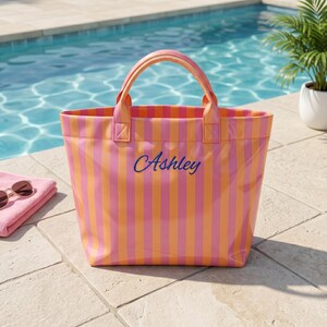 May include: A tote bag featuring vertical stripes in alternating shades of pink and orange. The bag has two matching handles and the name Ashley is embroidered in blue script lettering across the center of the front panel of the bag.