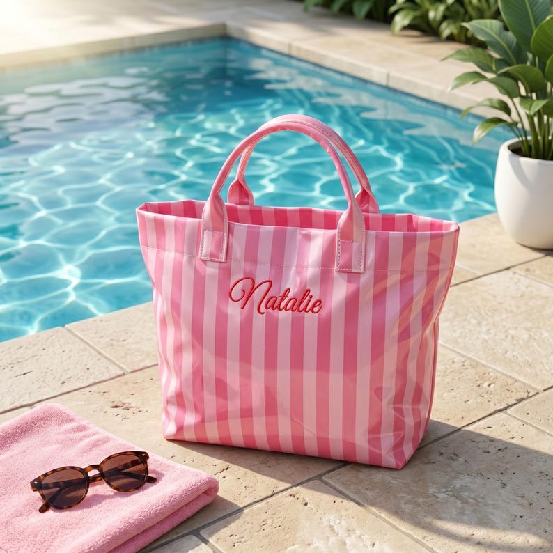 May include: A pink and white vertically striped tote bag with two top handles. The name Natalie is embroidered in red cursive script on the front center of the bag. The bag is positioned near a pink towel and brown sunglasses.