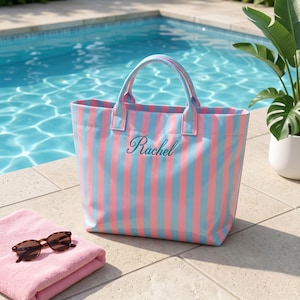 May include: A tote bag featuring vertical light blue and pink stripes with the name Rachel embroidered in dark teal script on the front. The bag has two matching striped handles and a rectangular base, positioned near a pink towel and sunglasses.
