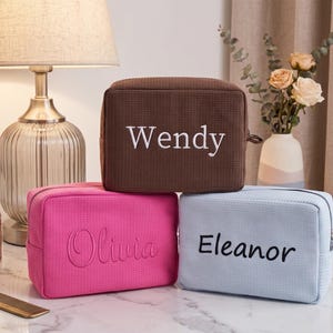 Puede incluir: Tres neceseres rectangulares de tela con textura de nido de abeja. El neceser marrón superior lleva el nombre Wendy bordado en blanco. El neceser rosa de abajo a la izquierda tiene el nombre Olivia bordado en rosa, y el azul claro de abajo a la derecha tiene el nombre Eleanor bordado en negro.