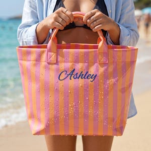 May include: A rectangular tote bag featuring vertical stripes in alternating shades of bright pink and orange. The bag has two top handles and the name Ashley embroidered in blue script lettering on the front. The surface is covered in water droplets.