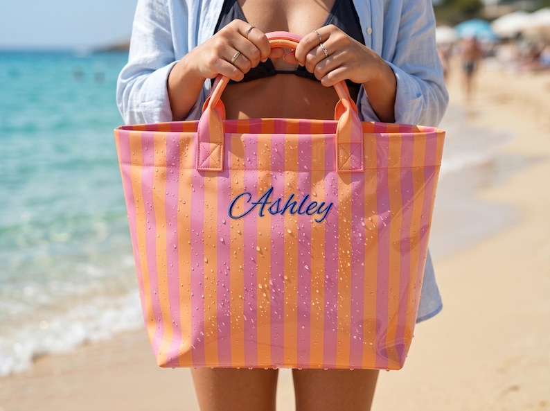 May include: A rectangular tote bag featuring vertical stripes in alternating shades of bright pink and orange. The bag has two top handles and the name Ashley embroidered in blue script lettering on the front. The surface is covered in water droplets.