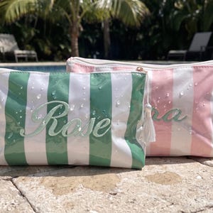 May include: Two rectangular zippered pouches with vertical stripes. One pouch is green and white with the embroidered name Rose, and the other is pink and white with an embroidered name. Both feature a small tassel and water droplets on the surface.