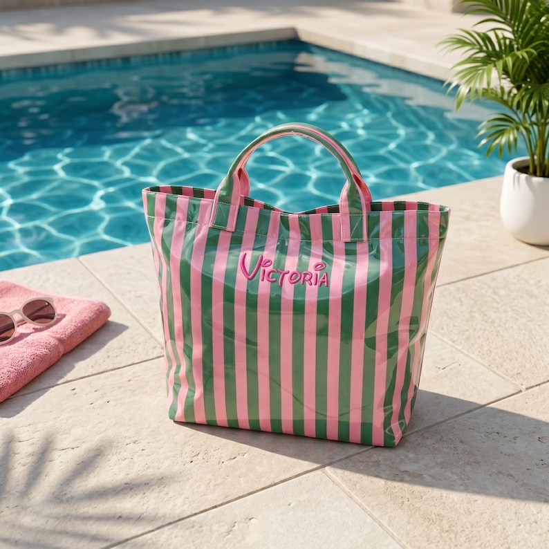 May include: A tote bag featuring vertical pink and dark green stripes with two top handles. The name Victoria is embroidered in pink script on the front center of the bag. The material has a glossy finish and a structured rectangular shape.