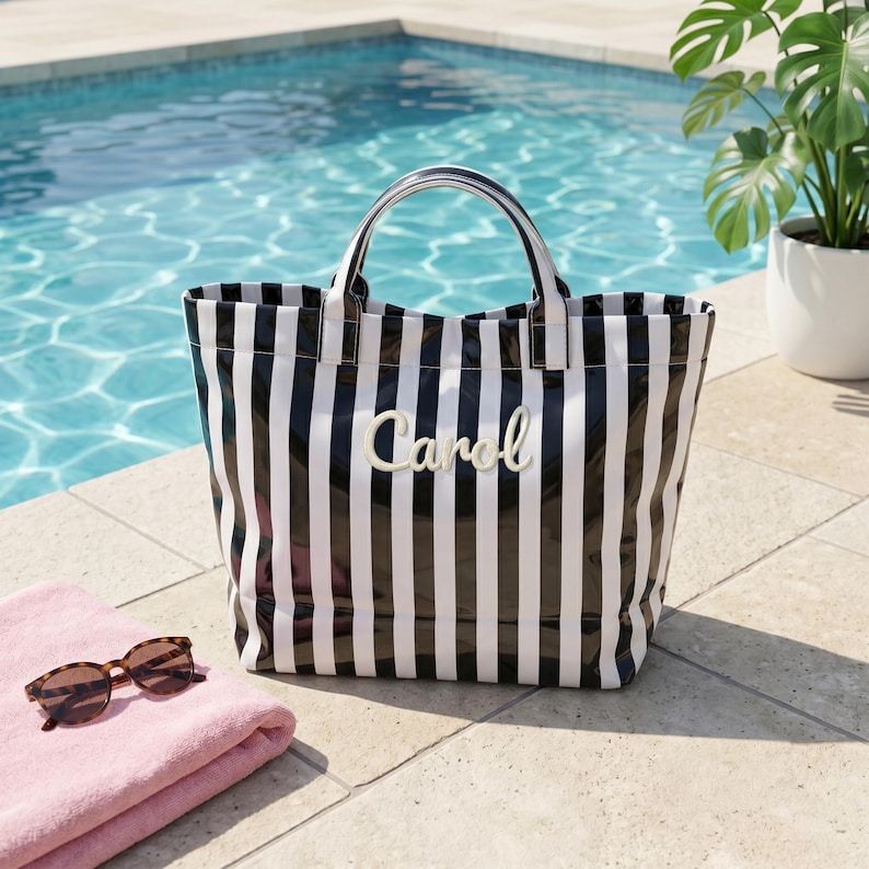 May include: A black and white vertical striped tote bag featuring two top handles and the name Carol written in a white script font on the front center. The bag has a structured rectangular shape and a glossy finish, positioned on a tiled surface.