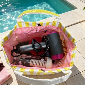 May include: A yellow and white striped tote bag with a pink interior lining. The bag contains a black and magenta hair dryer, a clear water bottle with a white filter insert, and a light-colored hair brush. The bag features white handles.