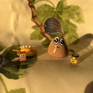May include: A whimsical still life featuring a hand-painted, brown, egg-shaped character with large eyes. Miniature food items, including a cake, a box of cookies, and a jar of honey, are arranged around it. The background is a fabric with a fig and leaf pattern.