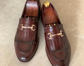 Mocasines de cuero marrón hechos a mano, con detalle de flecos y adornos metálicos, sin cordones.