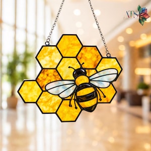 May include: Stained glass honeycomb with a black-outlined bee. The bee has yellow and black stripes, with white wings. The honeycomb is yellow and orange, with a black border. The piece hangs from a silver chain. The logo "ATS GLASS PATTERN" is in the upper right corner.