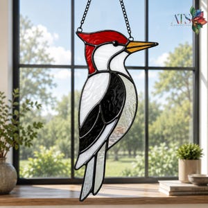 Op de afbeelding: Een glas-in-lood zonnevanger in de vorm van een specht met een rode kuif, wit gezicht en een zwart-wit lichaam. De vogel heeft een gele snavel en hangt aan een zwarte metalen ketting. De glasstukken zijn verbonden door donkere metalen soldeerlijnen.