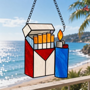 May include: Stained glass art piece depicting a cigarette pack and a lighter. The pack is red, white, and blue, with cigarettes visible inside. A blue lighter with a flame is next to the pack. The piece hangs from a chain.