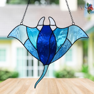 Puede incluir: Atrapasoles de raya manta de vidriera en tonos azules, con una cadena negra para colgar. El diseño presenta un cuerpo central azul oscuro, rodeado de alas y acentos azules más claros. Una pieza decorativa para ventanas o paredes.