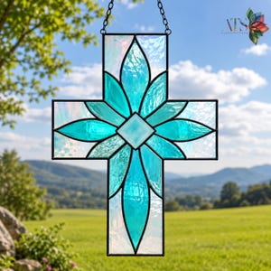 May include: Stained glass cross featuring a turquoise floral design. The cross is made of iridescent glass with a black border and a chain for hanging. The background shows a green field, trees, and a blue sky.