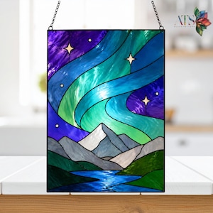 Op de afbeelding: Gebrandschilderd glaspaneel met een aurora borealis-sc&egrave;ne. Het kunstwerk toont een levendige weergave van blauw, groen en paars, met witte bergtoppen en een stromende rivier. Het paneel is zwart omlijst en opgehangen aan een ketting.