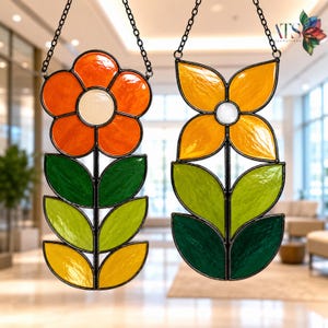 Puede incluir: Dos atrapasoles de vidrieras en forma de flor que cuelgan de cadenas negras. La flor de la izquierda tiene pétalos naranjas y un centro crema con hojas verdes y amarillas. La flor de la derecha tiene pétalos amarillos y un centro blanco.