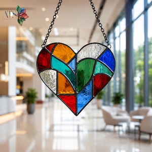 May include: A heart-shaped stained glass suncatcher featuring a geometric pattern of multicolored glass segments in red, orange, yellow, green, blue, and clear textured white, all held together by dark metal lead lines and suspended by a black chain.