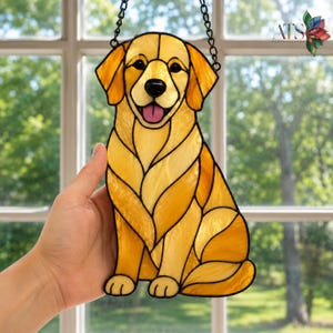May include: Stained glass window decoration of a golden retriever dog. The dog is rendered in shades of yellow and gold with black outlines, a pink tongue, and a black chain for hanging. The artwork is held in a hand.