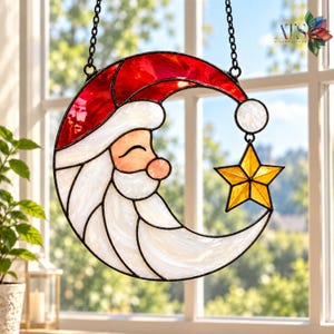 May include: A stained glass suncatcher shaped like a crescent moon featuring a Santa Claus face with a red hat, white beard, and a yellow star hanging from the tip of the hat. The piece is constructed with black metal lead lines and a chain.