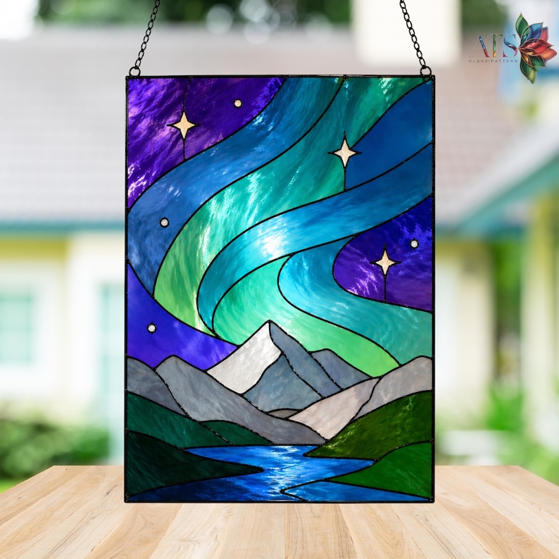 Op de afbeelding: Gebrandschilderd glaspaneel met een nachtsc&egrave;ne met het noorderlicht. Het kunstwerk toont wervelende banden van blauw, groen en paars licht, met witte bergen en een blauwe rivier op de voorgrond. Inclusief ophangketting.