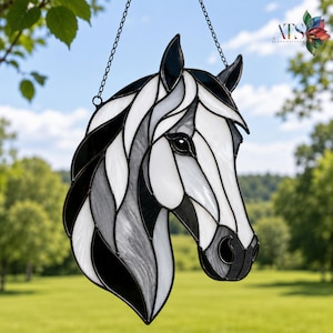 May include: Stained glass horse head suncatcher in black, white, and grey. The horse head is hanging from a chain. The suncatcher is in front of a green background with trees and a blue sky.